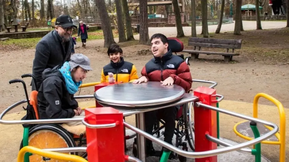 Zwei junge Menschen sind mit ihren Rollstühlen auf das Karussell im Seehofpark gefahren, daneben stehen ein Kind und ein Erwachsener.