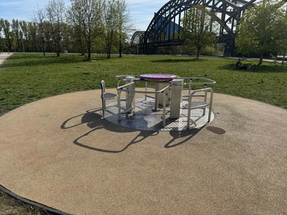 Das Karussell im Hafenpark mit Haltebügeln und Sitzen auf kreisrundem Fallschutzbelag.